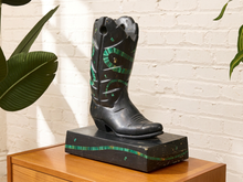 Load image into Gallery viewer, A black cowboy boot sculpture with green inlay details, displayed on a matching black rectangular base on a wooden table against a white brick wall with green leaves in the foreground.