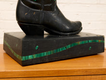 Load image into Gallery viewer, A black wooden sculpture of a cowboy boot mounted on a matching rectangular base, featuring green inlaid accents, displayed on a wooden table against a white brick wall.