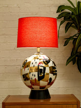 Load image into Gallery viewer, A table lamp with a bright red fabric drum shade and a glossy, abstract-patterned ceramic base in gold, white, black, and red, sitting on a wooden side table against a white brick wall with a green plant in the background.