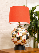 Load image into Gallery viewer, A decorative table lamp with a bright orange-red drum shade and a glossy, patterned spherical base featuring gold, black, white, and red geometric shapes, sitting on a wooden table against a white brick wall with a green plant in the background.