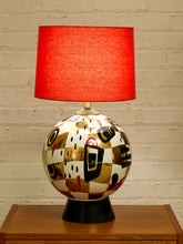 Load image into Gallery viewer, A table lamp with a bright red fabric drum shade and a spherical ceramic base featuring an abstract geometric pattern in gold, black, white, and red, sitting on a wooden surface against a white brick wall.