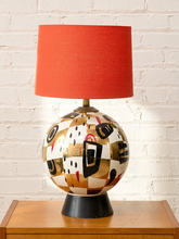 Load image into Gallery viewer, A mid-century modern table lamp featuring a glossy, abstract geometric patterned globe base in gold, black, white, and red, with a solid orange-red fabric drum shade, sitting on a wooden side table against a white brick wall.