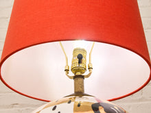 Load image into Gallery viewer, A close-up of a table lamp featuring a vibrant red fabric drum shade, a brass-colored socket, and a decorative ceramic base with a black and white pattern, set against a white brick wall.