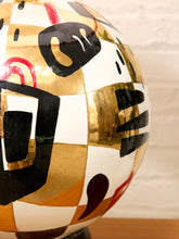 Load image into Gallery viewer, A close-up of a decorative spherical object featuring an abstract geometric pattern with black, gold, white, and red painted sections, set against a light brick wall background.