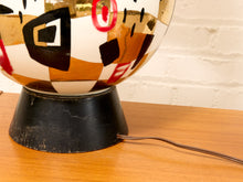 Load image into Gallery viewer, A close-up of a vintage mid-century modern table lamp featuring a glossy, hand-painted glass globe with abstract geometric patterns in black, red, brown, and white, resting on a black metal base on a wooden table.