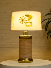 Load image into Gallery viewer, A mid-century modern table lamp with a glowing cream fabric shade featuring gold abstract shapes, and a cylindrical striped base in beige and brown with gold metal trim, set on a white round table against a white brick wall.