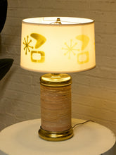 Load image into Gallery viewer, A mid-century modern table lamp with a ribbed metallic gold base and a white drum shade decorated with yellow retro geometric starburst patterns, shown lit on a white round table against a white brick wall.