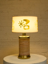 Load image into Gallery viewer, A mid-century modern table lamp with a cylindrical striped base and a drum shade decorated with gold mid-century geometric and starburst motifs, shown lit against a warm brick wall.