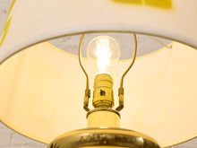 Load image into Gallery viewer, Close-up of a brass lamp base and glowing light bulb under a cream-colored fabric lampshade.
