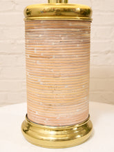 Load image into Gallery viewer, A cylindrical table lamp featuring a textured, ribbed beige and white striped body, finished with polished gold brass top and bottom bases, shown on a white surface against a white brick wall.