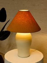 Load image into Gallery viewer, A table lamp with a textured brown fabric cone shade and a cream-to-white ceramic base, illuminated with warm light against a white brick wall.
