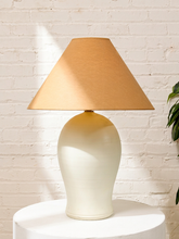 Load image into Gallery viewer, A table lamp with a tapered beige fabric shade and a glossy cream ceramic base, displayed on a white round pedestal against a white brick wall.