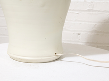 Load image into Gallery viewer, Close-up of the base of a cream-colored ceramic table lamp with a visible power cord exiting the side, set against a white brick wall background.