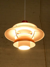 Load image into Gallery viewer, A modern multi-tiered pendant light with a warm glowing white glass shade and a red top cap, hanging against a textured beige brick wall.