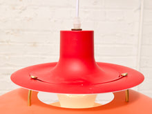 Load image into Gallery viewer, A close-up of a mid-century modern pendant light with a vibrant red conical shade, a wide red flared rim, and small brass support legs, set against a white brick wall background.