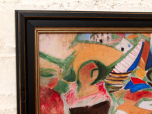 Load image into Gallery viewer, A close-up of a framed abstract painting featuring bold brushstrokes in orange, green, red, blue, and cream, set in a black frame with a gold inner trim against a textured off-white brick wall.