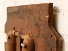 Load image into Gallery viewer, A close-up of a rustic, weathered brown wooden wall-mounted rack with curved hooks, shown against a white painted brick wall.