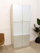 Load image into Gallery viewer, A tall, narrow modern cabinet with four doors, featuring a cream-colored frame and doors with white and light beige panels, set against a white brick wall with a plant and chair nearby.