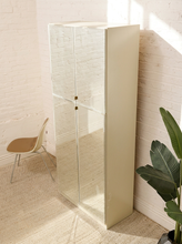 Load image into Gallery viewer, A tall, narrow cream-colored cabinet with mirrored doors stands in a bright room with white brick walls, a beige chair, and a green potted plant.