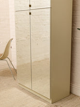 Load image into Gallery viewer, A tall, narrow cabinet with mirrored doors and a solid beige side panel, shown in a room setting with a white brick wall, beige rug, and a partial view of a chair.