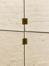 Load image into Gallery viewer, Close-up of a modern glass cabinet or door with two brass-colored metal clamps holding the glass panels together, set against a light beige brick wall background.