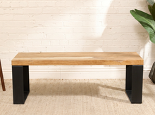 Load image into Gallery viewer, A modern industrial bench with a light natural wood top and black metal block legs, set against a white brick wall and beige rug.