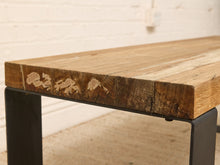 Load image into Gallery viewer, A close-up of a rustic wooden table with a thick, weathered oak top showing natural grain and knots, supported by a black metal frame leg.