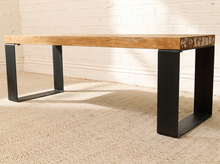 Load image into Gallery viewer, A modern industrial-style coffee table with a thick, live-edge natural oak wood top and matte black U-shaped metal legs, set on a beige textured rug against a white brick wall.