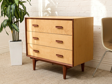 Load image into Gallery viewer, A mid-century modern three-drawer dresser with a light natural wood finish and dark brown trim, shown in a styled room setting next to a potted plant and a chair.