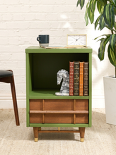 Load image into Gallery viewer, A mid-century modern side table with a sage green painted frame, a wooden drawer front, and tapered wood legs, styled with books, a chess piece, and a mug against a white brick wall.