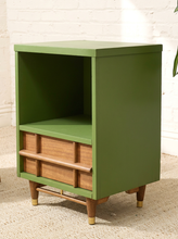 Load image into Gallery viewer, A mid-century modern side cabinet with a sage green painted finish and natural wood drawer fronts, legs, and footrest, set against a white brick wall and light beige rug.