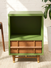 Load image into Gallery viewer, A mid-century modern open storage cabinet with a sage green painted frame and drawer front, paired with natural wood legs and drawer pulls, set against a white brick wall background.