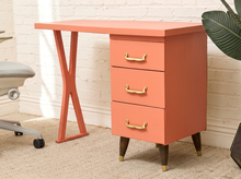 Load image into Gallery viewer, A coral pink three-drawer storage cabinet with gold handles and dark legs, styled next to a matching coral desk in a bright room with a white brick wall and a plant.