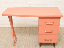 Load image into Gallery viewer, A mid-century modern desk in a warm coral pink color, featuring three drawers with gold handles and tapered legs, set against a white brick wall and beige carpet.