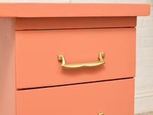 Load image into Gallery viewer, A close-up of a coral pink wooden drawer with a curved, polished brass pull handle, set against a white brick wall background.