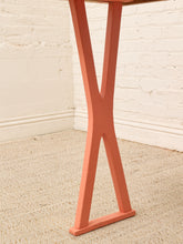 Load image into Gallery viewer, A close-up of a table leg in a terracotta or salmon pink color, standing on a beige textured rug against a white painted brick wall.