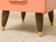 Load image into Gallery viewer, Close-up of a mid-century modern side table or stool with a coral pink painted frame, dark brown tapered wooden legs, and brass metal feet, set on a textured beige woven rug.