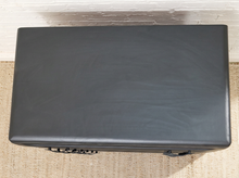 Load image into Gallery viewer, A black hard-sided rectangular case or briefcase with metal latches, photographed from above on a beige woven rug against a white brick wall.