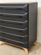 Load image into Gallery viewer, A mid-century modern 7-drawer chest of drawers in matte black with light natural wood legs, shown against a white brick wall and woven rug.
