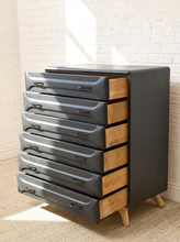 Load image into Gallery viewer, A mid-century modern 6-drawer chest of drawers with dark gray metal drawer fronts, natural wood sides, and tapered wooden legs, set against a white brick wall.
