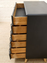 Load image into Gallery viewer, A mid-century modern chest of drawers with a black leather-look finish and five open oak wood drawers, set against a white brick wall and beige carpet.