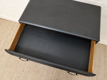 Load image into Gallery viewer, A black upholstered storage drawer with a matching lid, shown open to reveal a black interior lining and natural wood drawer sides, set on a beige rug against a white brick wall.