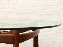Load image into Gallery viewer, Close-up of a round glass-top table with a curved medium-brown wooden frame and legs, set against a white painted brick wall.
