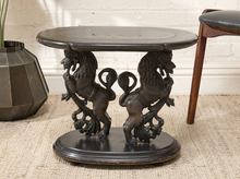 Load image into Gallery viewer, A dark carved wooden and metal side table featuring two lion figures supporting the base, set on a woven rug with a white brick wall and a vase in the background.