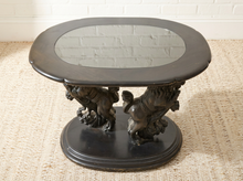 Load image into Gallery viewer, A dark carved wooden end table with an oval mirror top and ornate sculptural legs, set on a woven rug against a white brick wall.