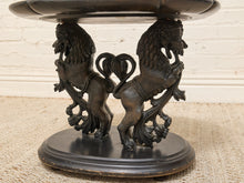 Load image into Gallery viewer, An ornate dark metal pedestal table base featuring two lion figures supporting a heart-shaped center motif, with a round dark top and base, photographed against a light brick wall and burlap surface.