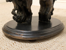 Load image into Gallery viewer, Close-up of the base of a dark bronze figurative sculpture, showing the feet of two animals standing on a round pedestal over a woven beige rug.