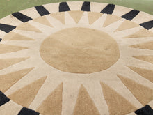 Load image into Gallery viewer, A round coir rug with a sunburst pattern in natural tan and beige, featuring alternating black and beige rays and a black outer border, shown on a green outdoor surface.