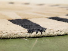 Load image into Gallery viewer, Close-up of a woven rug corner showing a beige base with bold black geometric stripes and a frayed natural-fiber edge, photographed over a green floor.