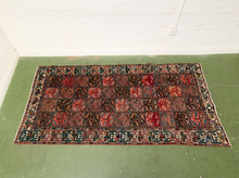 Load image into Gallery viewer, A rectangular Persian-style area rug with a complex geometric patchwork pattern in red, brown, black, and beige, displayed on a green floor against a white brick wall.